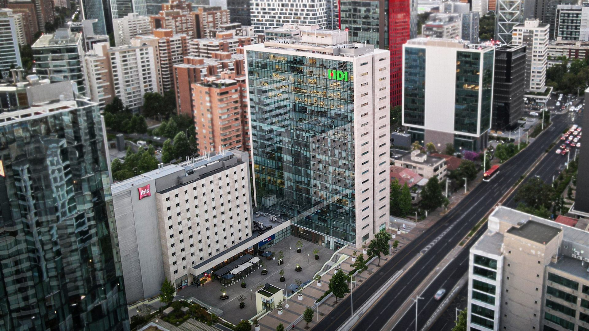 Vista aérea al atardecer de la torre de vidrio y piedra junto al Hotel IBIS, con su plaza de acceso sobre Av. Manquehue Norte